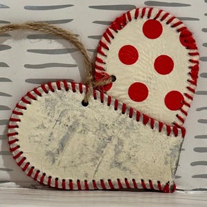 May include: A white heart-shaped ornament with red stitching and red polka dots. The ornament has a twine loop for hanging.