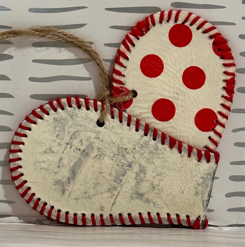 May include: A white heart-shaped ornament with red stitching and red polka dots. The ornament has a twine loop for hanging.