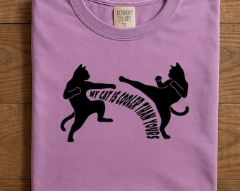 Könnte beinhalten: Ein lilafarbenes T-Shirt mit Rundhalsausschnitt und einem schwarzen Aufdruck von zwei Katzen in Karate-Posen. Der Text "MY CAT IS COOLER THAN YOURS" ist in einem Bogen gedruckt. Das Shirt hat ein "Comfort Colors"-Etikett.