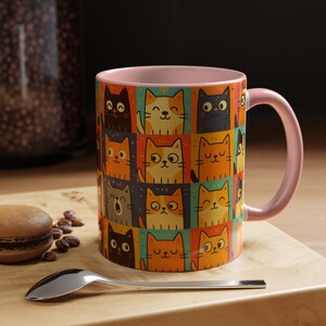 Puede incluir: Una taza de café de cerámica con interior y asa rosas. El exterior presenta un patrón repetido de gatos de dibujos animados en varios colores y expresiones, sobre un fondo colorido. La taza está sobre una superficie de madera.