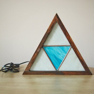 K&ouml;nnte beinhalten: Eine dreieckige Holzlampe mit einem Buntglasdesign. Das Glas ist wei&szlig; mit einem blauen Dreieck in der Mitte.