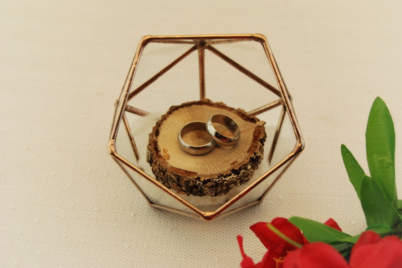 May include: Two gold wedding bands on a wooden ring bearer pillow inside a geometric glass box.