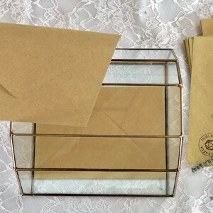 May include: A glass box with a brown envelope inside. The box has a gold metal frame and is sitting on a white lace tablecloth. The envelope has a brown paper texture and is folded in half. There is a brown envelope with a stamp that says "Mudita Arts & Crafts" on the right side of the image.
