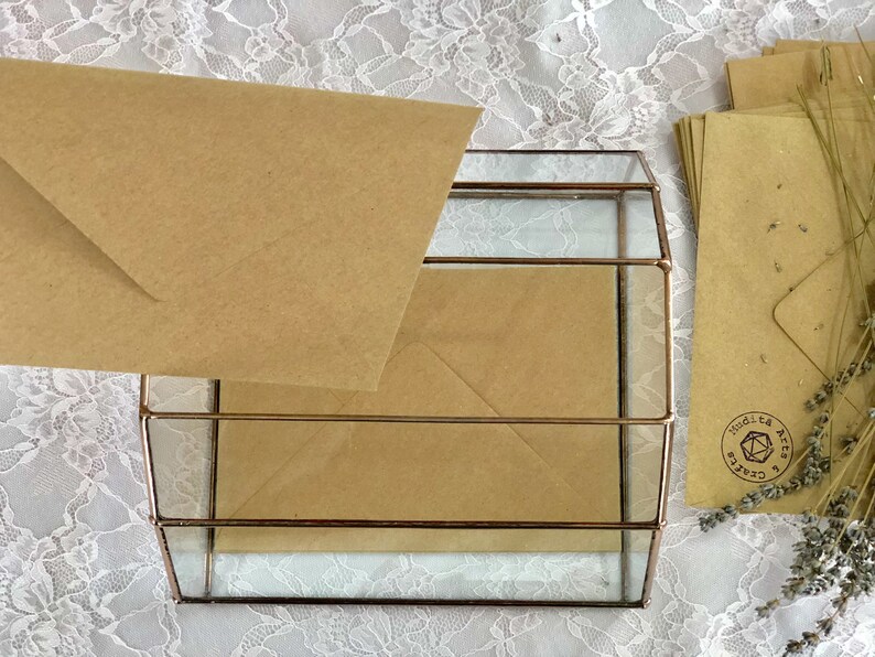 May include: A glass box with a brown envelope inside. The box has a gold metal frame and is sitting on a white lace tablecloth. The envelope has a brown paper texture and is folded in half. There is a brown envelope with a stamp that says "Mudita Arts & Crafts" on the right side of the image.
