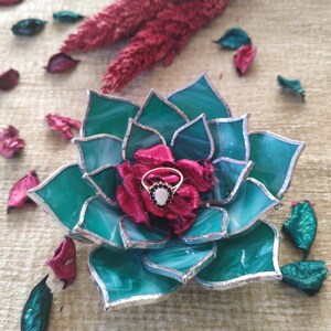 May include: A green glass succulent ring holder with a silver trim. A ring with a white stone and black accents is displayed in the center of the holder.