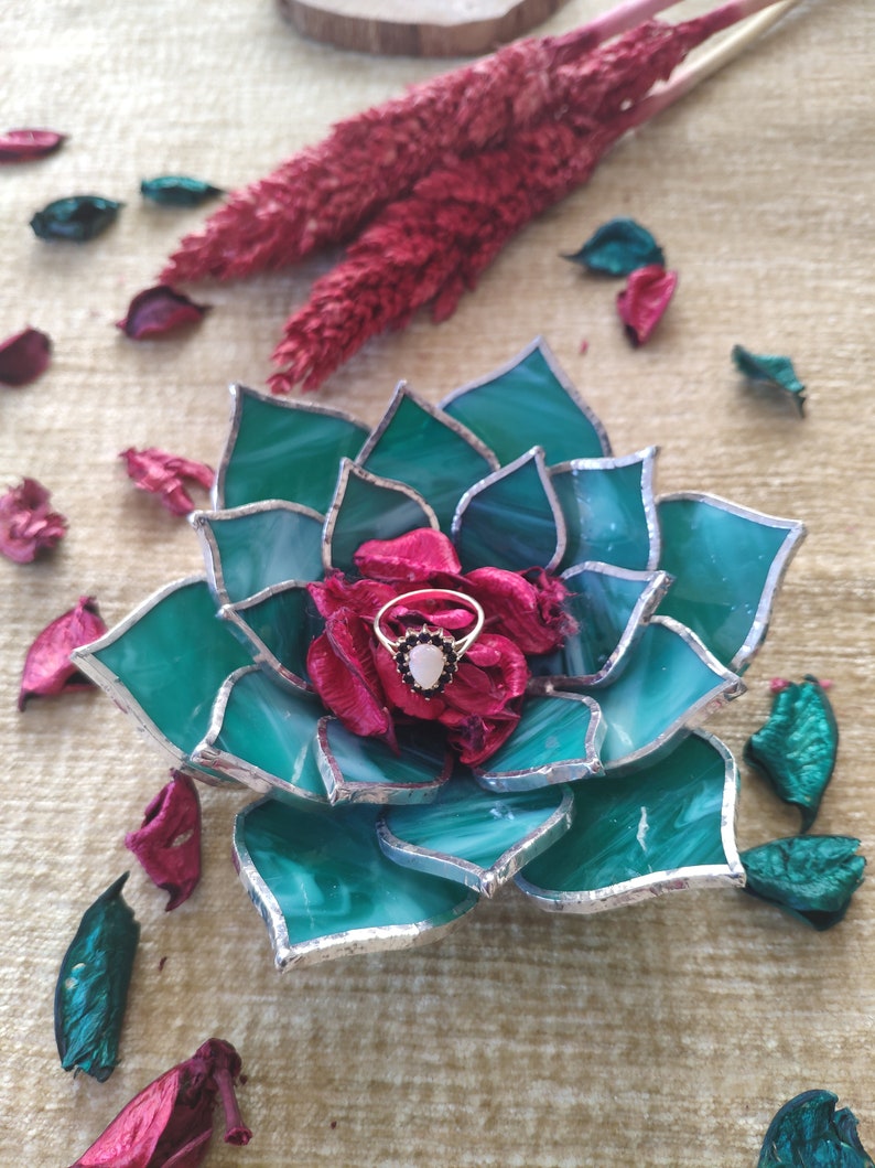 May include: A green glass succulent ring holder with a silver trim. A ring with a white stone and black accents is displayed in the center of the holder.