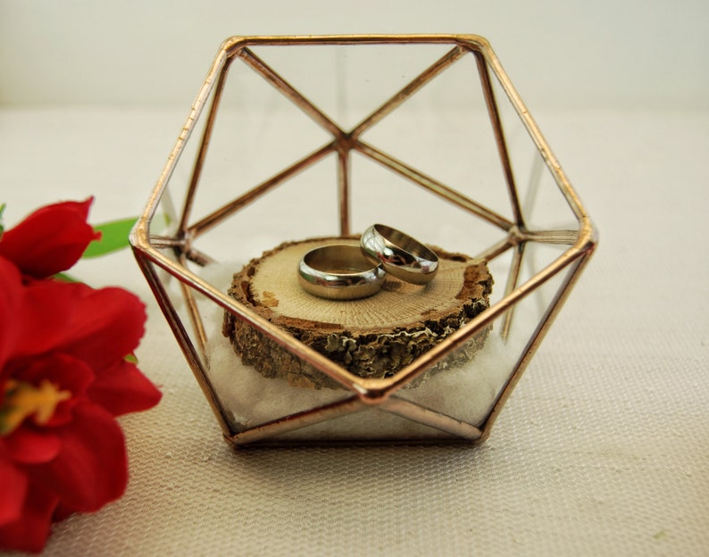 May include: Two silver wedding bands are displayed inside a geometric glass box with a copper frame. The rings are resting on a small wooden slice.