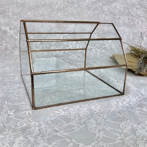 May include: A glass box with a copper frame, shaped like a house. The box is empty and sitting on a white lace tablecloth.