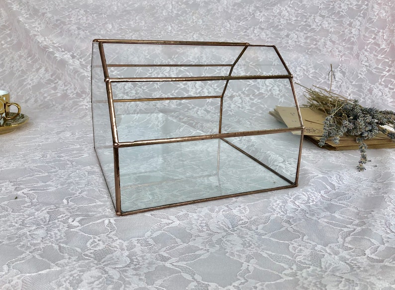 May include: A glass box with a copper frame, shaped like a house. The box is empty and sitting on a white lace tablecloth.
