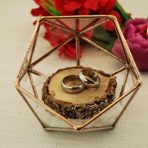 May include: Two gold wedding bands on a wooden slice inside a geometric glass box with a copper frame.