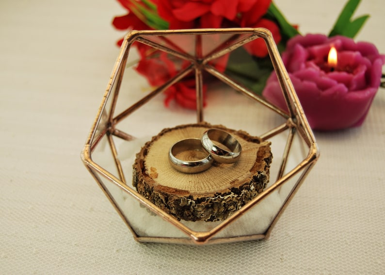 May include: Two gold wedding bands on a wooden slice inside a geometric glass box with a copper frame.