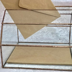 May include: A glass box with a copper frame, used for holding envelopes. The box has two levels and is filled with brown envelopes.
