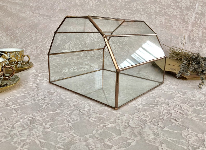 May include: A glass geometric box with a copper frame. The box is sitting on a white lace tablecloth.