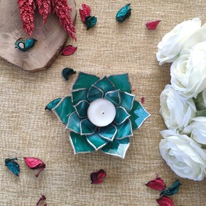 May include: Teal glass candle holder shaped like a flower with a white candle inside. The candle holder is surrounded by white flowers and red flower petals.