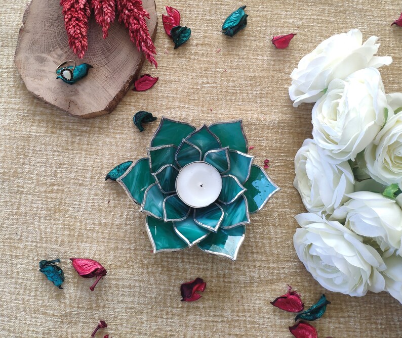 May include: Teal glass candle holder shaped like a flower with a white candle inside. The candle holder is surrounded by white flowers and red flower petals.