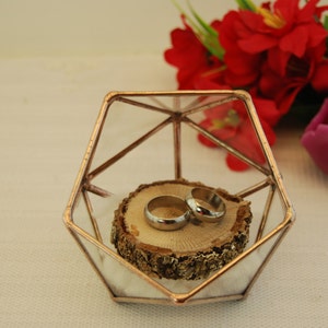 May include: Two gold wedding bands are displayed on a wooden slice inside a geometric glass and copper ring holder. The holder is on a white surface with red flowers and a pink candle in the background.