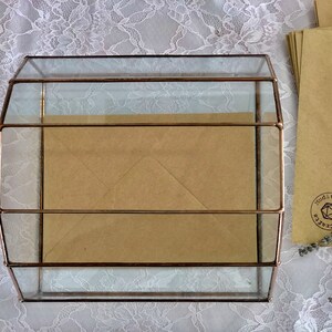 May include: A glass box with a copper frame, containing brown envelopes. The box is sitting on a white lace tablecloth.