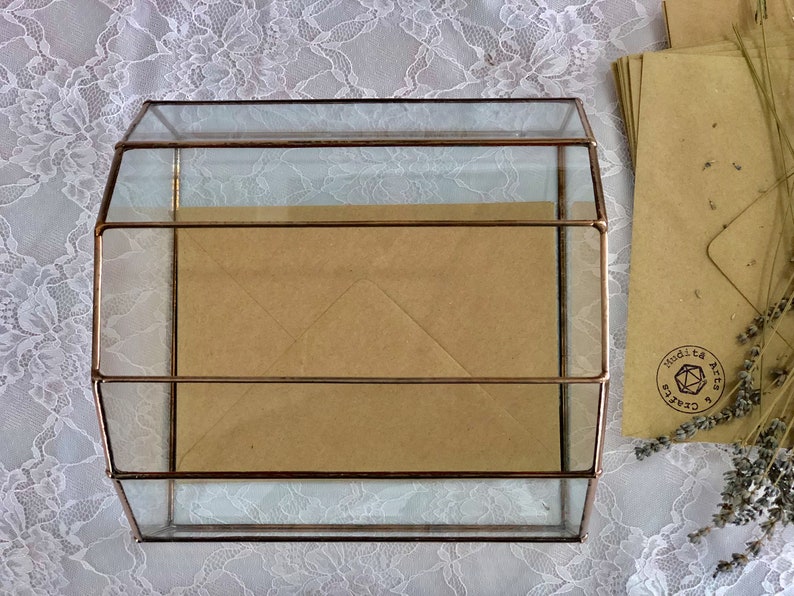 May include: A glass box with a copper frame, containing brown envelopes. The box is sitting on a white lace tablecloth.