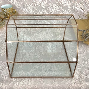 May include: A clear glass box with a copper frame. The box has three shelves and is sitting on a white lace tablecloth.
