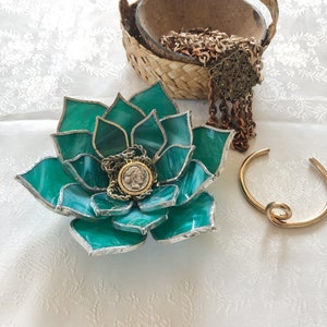 May include: A green glass succulent dish with a silver coin and gold chain inside. The dish is sitting on a white surface next to a woven basket and a gold bangle.