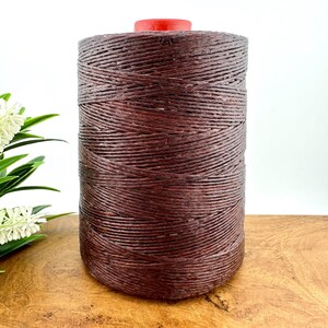 Puede incluir: Una bobina de hilo encerado marrón oscuro. El hilo está enrollado firmemente alrededor de la bobina, que tiene una parte superior roja.