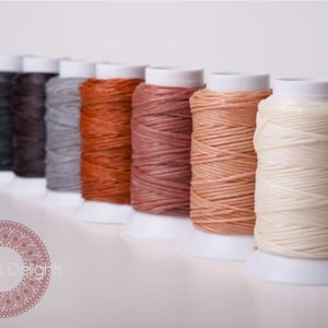 May include: A selection of thread spools in shades of black, brown, grey, orange, and cream. The spools are on white bases. The thread is tightly wound. The background is a soft, neutral colour.