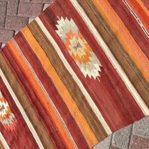 May include: A colorful, woven runner rug with a striped pattern in shades of red, orange, brown, and cream. The rug features two diamond-shaped, geometric designs in the center. The rug is made of a durable material and is suitable for high-traffic areas.