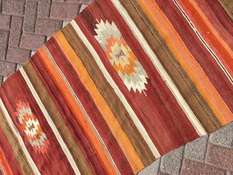 May include: A colorful, woven runner rug with a striped pattern in shades of red, orange, brown, and cream. The rug features two diamond-shaped, geometric designs in the center. The rug is made of a durable material and is suitable for high-traffic areas.