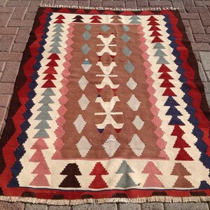May include: A rectangular, woven rug with a geometric pattern. The rug features a central brown field with white, pink, and blue diamond and tree-like shapes. The border is a mix of red, brown, and blue zigzags. The rug has a fringed edge.
