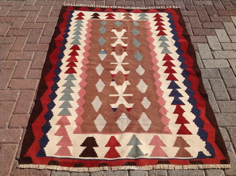 May include: A rectangular, woven rug with a geometric pattern. The rug features a central brown field with white, pink, and blue diamond and tree-like shapes. The border is a mix of red, brown, and blue zigzags. The rug has a fringed edge.