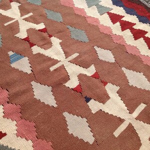 May include: A woven rug with a geometric pattern. The rug features a brown background with white, pink, red, blue, and gray diamond and cross-shaped designs. The rug's texture is visible, suggesting a handmade or artisanal quality.