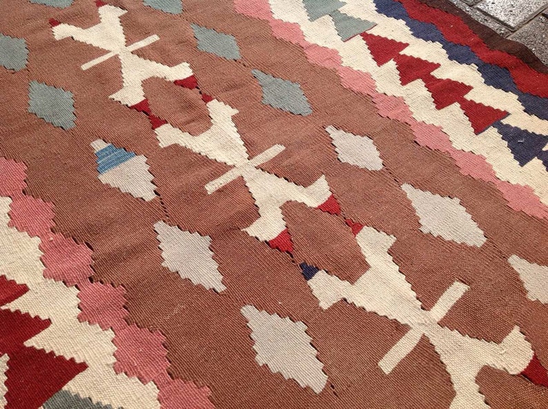 May include: A woven rug with a geometric pattern. The rug features a brown background with white, pink, red, blue, and gray diamond and cross-shaped designs. The rug's texture is visible, suggesting a handmade or artisanal quality.