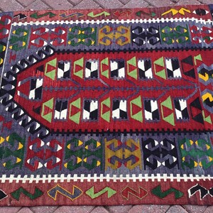 May include: A rectangular, hand-woven rug with a geometric pattern. The rug features a central red field with black, white, and green shapes. The border includes repeating patterns in shades of gray, green, yellow, and red. The rug has a fringed edge.