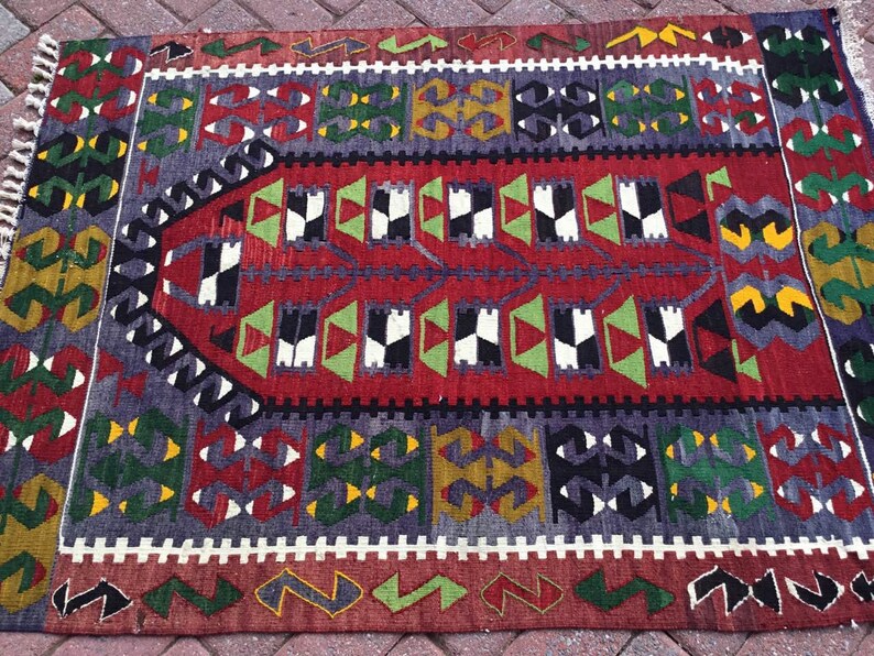 May include: A rectangular, hand-woven rug with a geometric pattern. The rug features a central red field with black, white, and green shapes. The border includes repeating patterns in shades of gray, green, yellow, and red. The rug has a fringed edge.