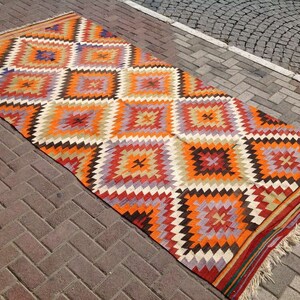 May include: A colorful, geometric patterned rug with a diamond design in shades of orange, red, brown, white, and purple. The rug has fringe on the edges.