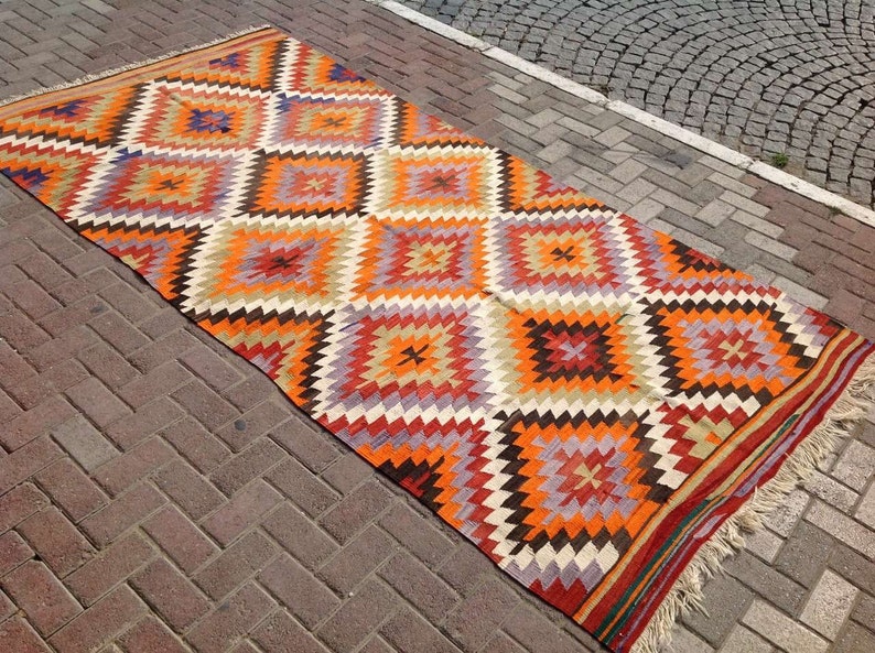 May include: A colorful, geometric patterned rug with a diamond design in shades of orange, red, brown, white, and purple. The rug has fringe on the edges.