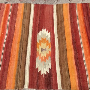 May include: A colorful, striped rug with a central geometric design. The rug features vertical stripes in shades of red, orange, brown, and cream. The central design is a stylized geometric pattern in orange, cream, and brown.