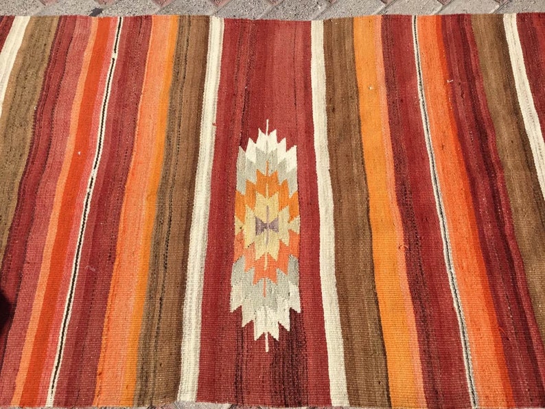 May include: A colorful, striped rug with a central geometric design. The rug features vertical stripes in shades of red, orange, brown, and cream. The central design is a stylized geometric pattern in orange, cream, and brown.