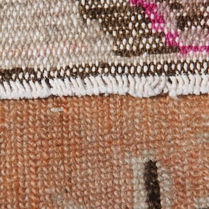 May include: Close-up of a section of a woven rug. The rug features a mix of colors, including brown, beige, and orange. The top portion has a pattern with white and pink accents, while the bottom is a solid orange with dark brown shapes.