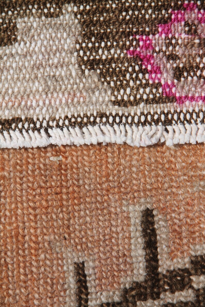 May include: Close-up of a section of a woven rug. The rug features a mix of colors, including brown, beige, and orange. The top portion has a pattern with white and pink accents, while the bottom is a solid orange with dark brown shapes.