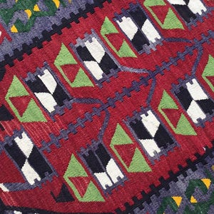 May include: Close-up of a red, geometric patterned rug. The rug features repeating patterns of black and white diamonds, green triangles, and blue and yellow accents. The rug has a textured appearance.