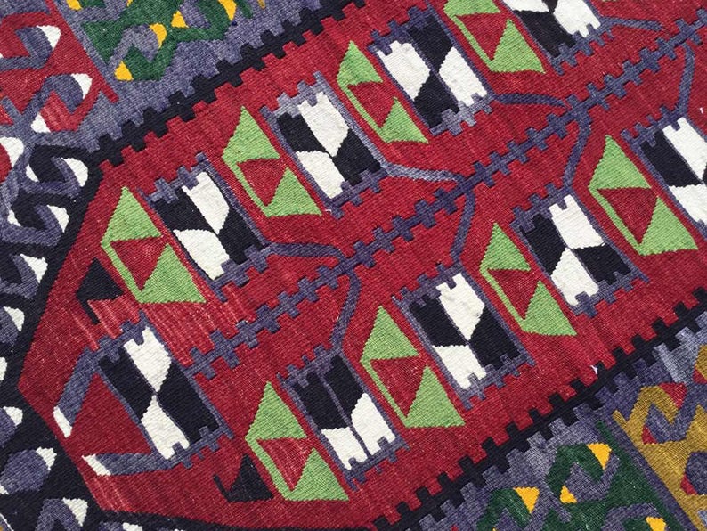 May include: Close-up of a red, geometric patterned rug. The rug features repeating patterns of black and white diamonds, green triangles, and blue and yellow accents. The rug has a textured appearance.