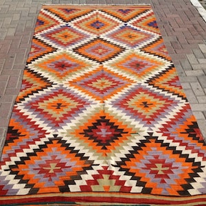 May include: A colorful, geometric rug with a diamond pattern in shades of red, orange, brown, white, and purple. The rug has a traditional, hand-woven look.