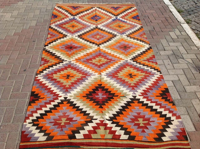 May include: A colorful, geometric rug with a diamond pattern in shades of red, orange, brown, white, and purple. The rug has a traditional, hand-woven look.