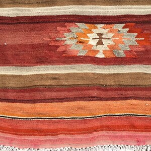 May include: A handwoven rug with a geometric design. The rug features horizontal stripes in shades of red, brown, orange, and cream. A central geometric pattern in orange, white, and gray adds a focal point. The rug has a fringed edge.