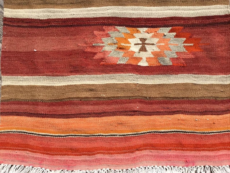 May include: A handwoven rug with a geometric design. The rug features horizontal stripes in shades of red, brown, orange, and cream. A central geometric pattern in orange, white, and gray adds a focal point. The rug has a fringed edge.