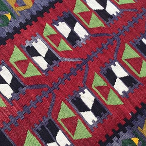 May include: Close-up of a woven rug with a geometric pattern. The rug features a red background with repeating black, white, and green shapes. Dark blue and yellow accents add contrast to the design. The rug's texture is visible.