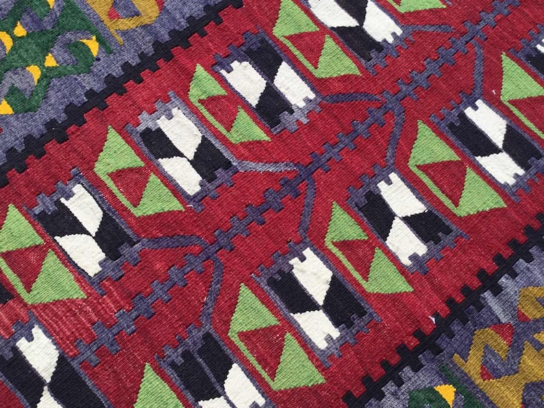 May include: Close-up of a woven rug with a geometric pattern. The rug features a red background with repeating black, white, and green shapes. Dark blue and yellow accents add contrast to the design. The rug's texture is visible.