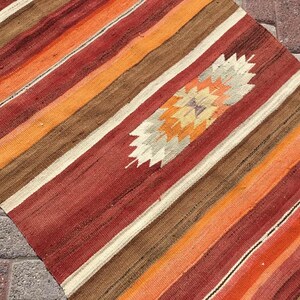 May include: A colorful, striped rug with a central diamond pattern. The rug features bands of red, orange, brown, and cream colors. The diamond pattern in the center has a starburst design with beige, orange, and purple hues.