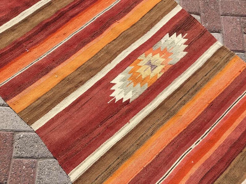 May include: A colorful, striped rug with a central diamond pattern. The rug features bands of red, orange, brown, and cream colors. The diamond pattern in the center has a starburst design with beige, orange, and purple hues.